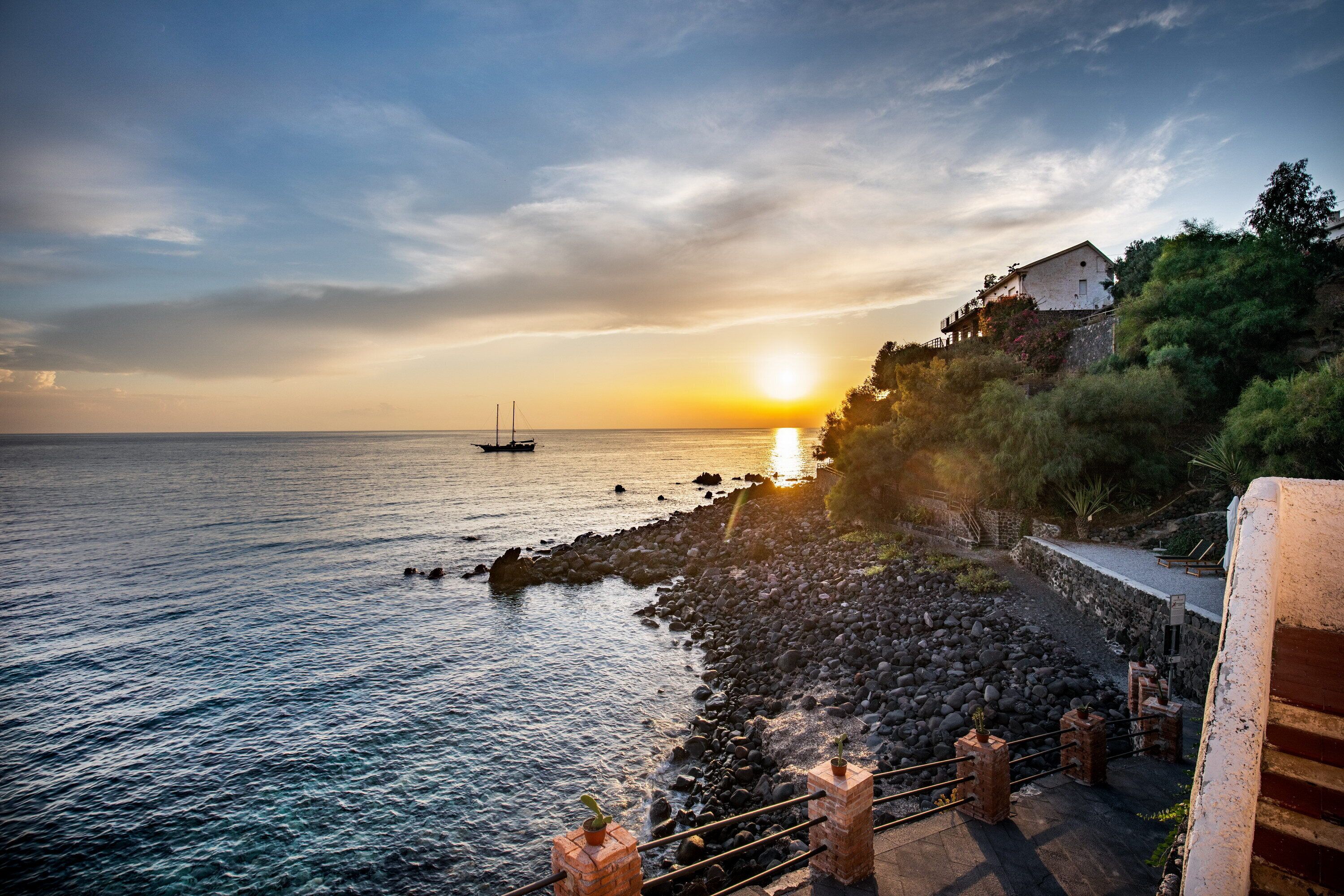 A coastal scene with a rocky shoreline, a building on a hill, and a boat in the water.