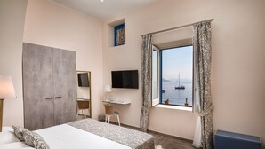 A hotel room with a bed, desk, TV, and a window with a view of the sea. - Hotel L’Ariana ISOLE EOLIE - UNA Esperienze (Leni)