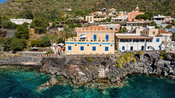 A coastal town with buildings along the shoreline, clear turquoise water, and rocky terrain. - Hotel L’Ariana ISOLE EOLIE - UNA Esperienze (Leni)