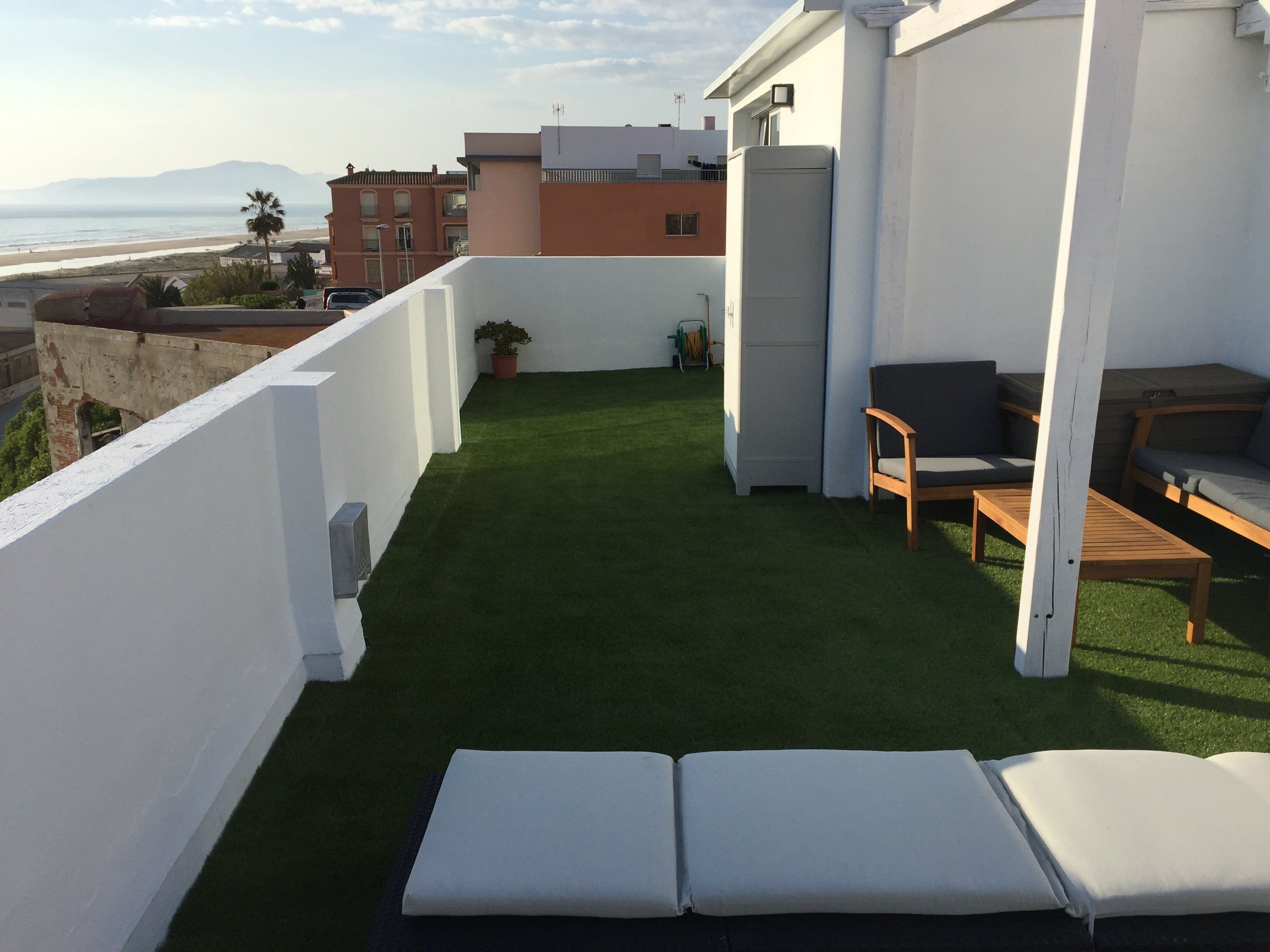 Eine Dachterrasse mit Kunstgrashalm, einer Sitzecke inklusive Tisch und Stühlen, und Blick auf das Meer sowie Gebäude im Hintergrund.