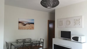 Un salon moderne avec une table à manger en verre, des chaises noires, un meuble de rangement blanc contenant un téléviseur et un micro-ondes, un élément décoratif mural et une porte donnant sur une autre pièce.
