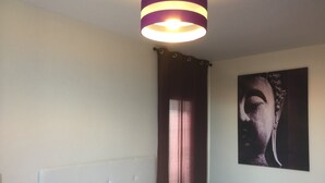 Une chambre à coucher comprenant un lit, une tête de lit, une lampe fixée au mur, une fenêtre avec des rideaux et un tableau.