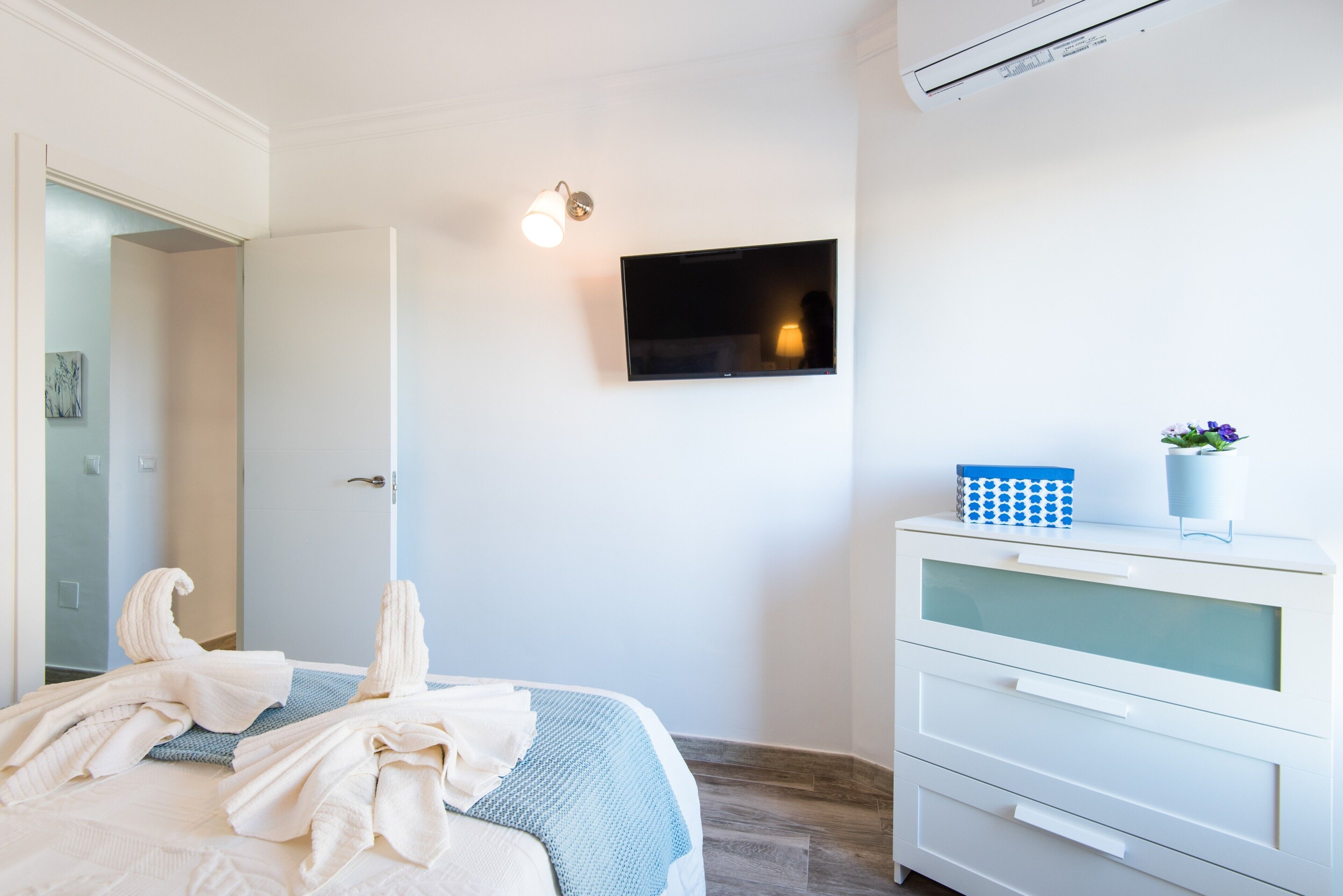 A hotel room with a bed, a TV mounted on the wall, a white dresser, and a door leading to another room.