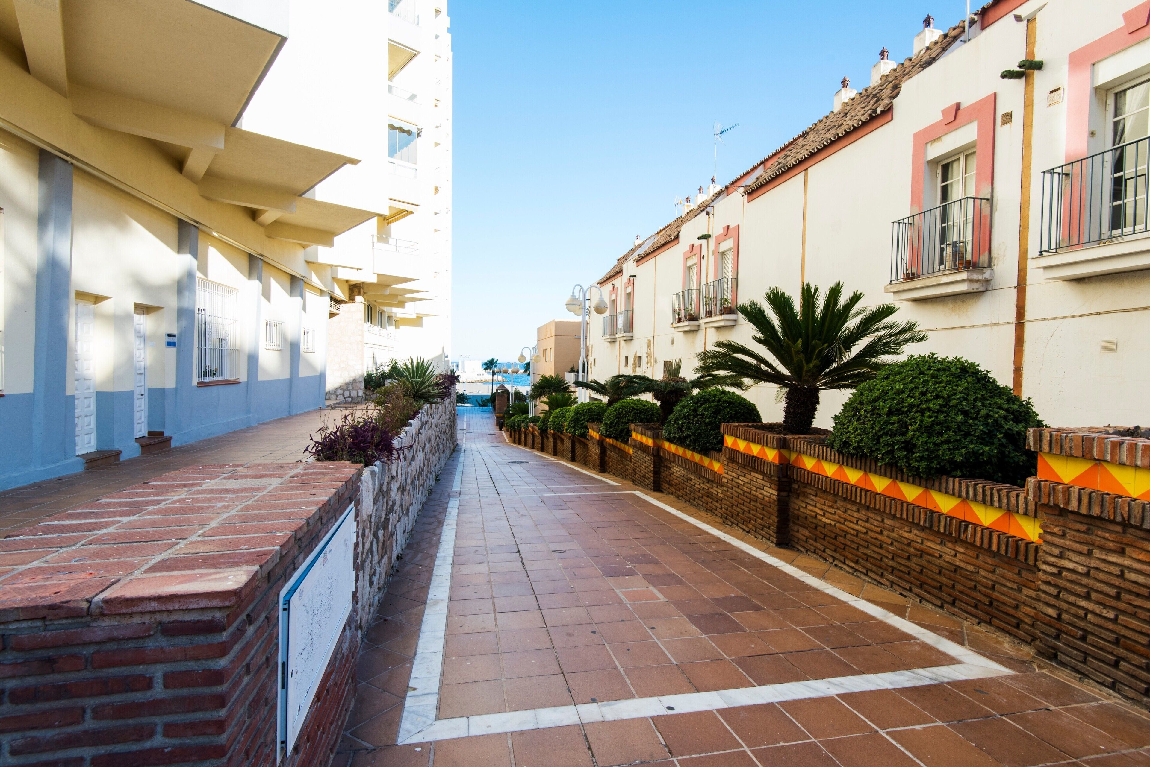 A narrow street with buildings on either side, featuring balconies and a brick wall with a decorative pattern.