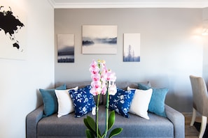 A grey sofa with floral cushions, a vase of pink orchids, and three framed artworks on the wall.