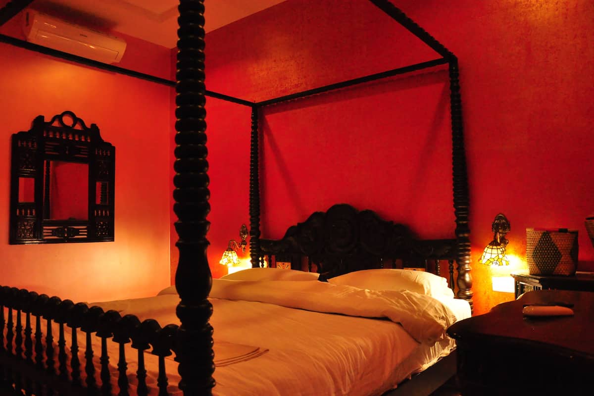 Una cama con dosel y cuatro columnas, una mesita de noche con lámpara, y un espejo fijado a la pared.