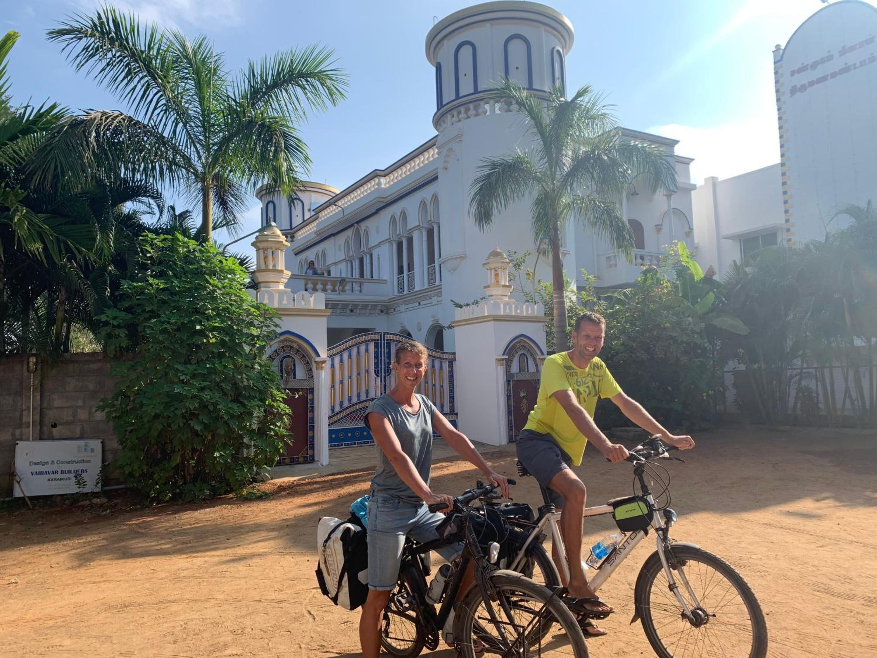 Two people on bicycles in front of a building with a white facade and arched windows.
