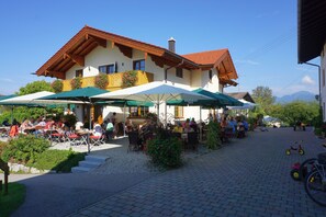 Relax in a shaded outdoor space with tables, chairs, and umbrellas, surrounded by a traditional building with a sloping roof and balcony railings.