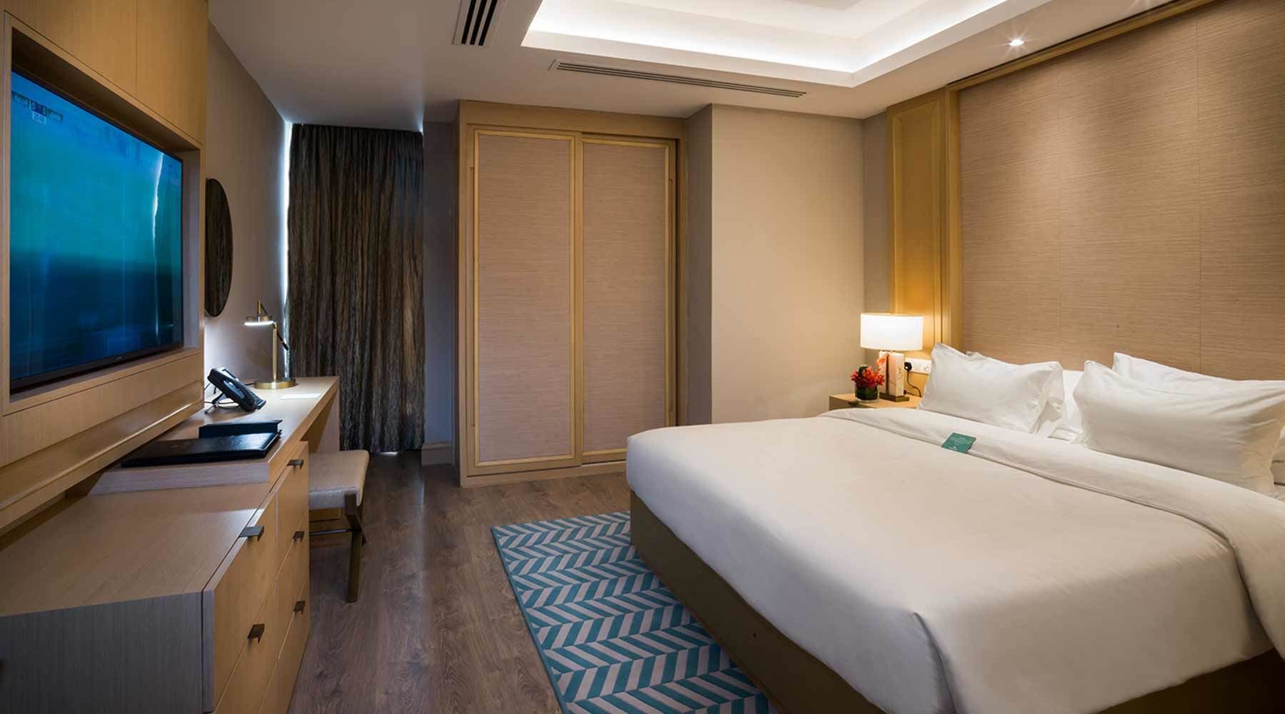 A hotel room with a bed, a desk with a computer, a TV, a wardrobe, and a nightstand with a lamp.