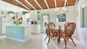 A kitchen with a central island, white cabinets, a dining area with wicker chairs, and a ceiling with exposed wooden beams. - Old Florida Surf Cottage (Jensen Beach)