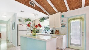 A kitchen with a central island, white cabinets, a window with a mesh screen, and a door with a green frame. - Old Florida Surf Cottage (Jensen Beach)