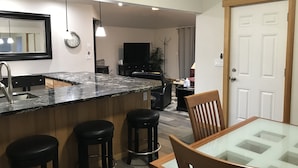 A modern kitchen with a marble countertop, a sink, and three stools. There is a dining area with a glass-top table and wooden chairs. - Direct Dune Access Sleeps 10 In House, 6 In BunkHouses - Bath Houses. (North Bend)
