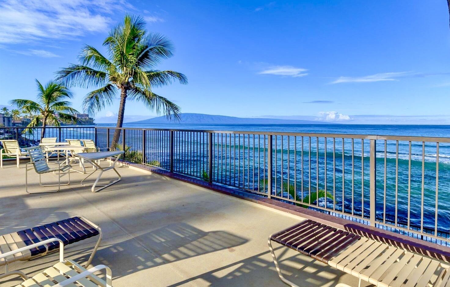 A balcony with lounge chairs, a table, and a view of the ocean and a mountain.