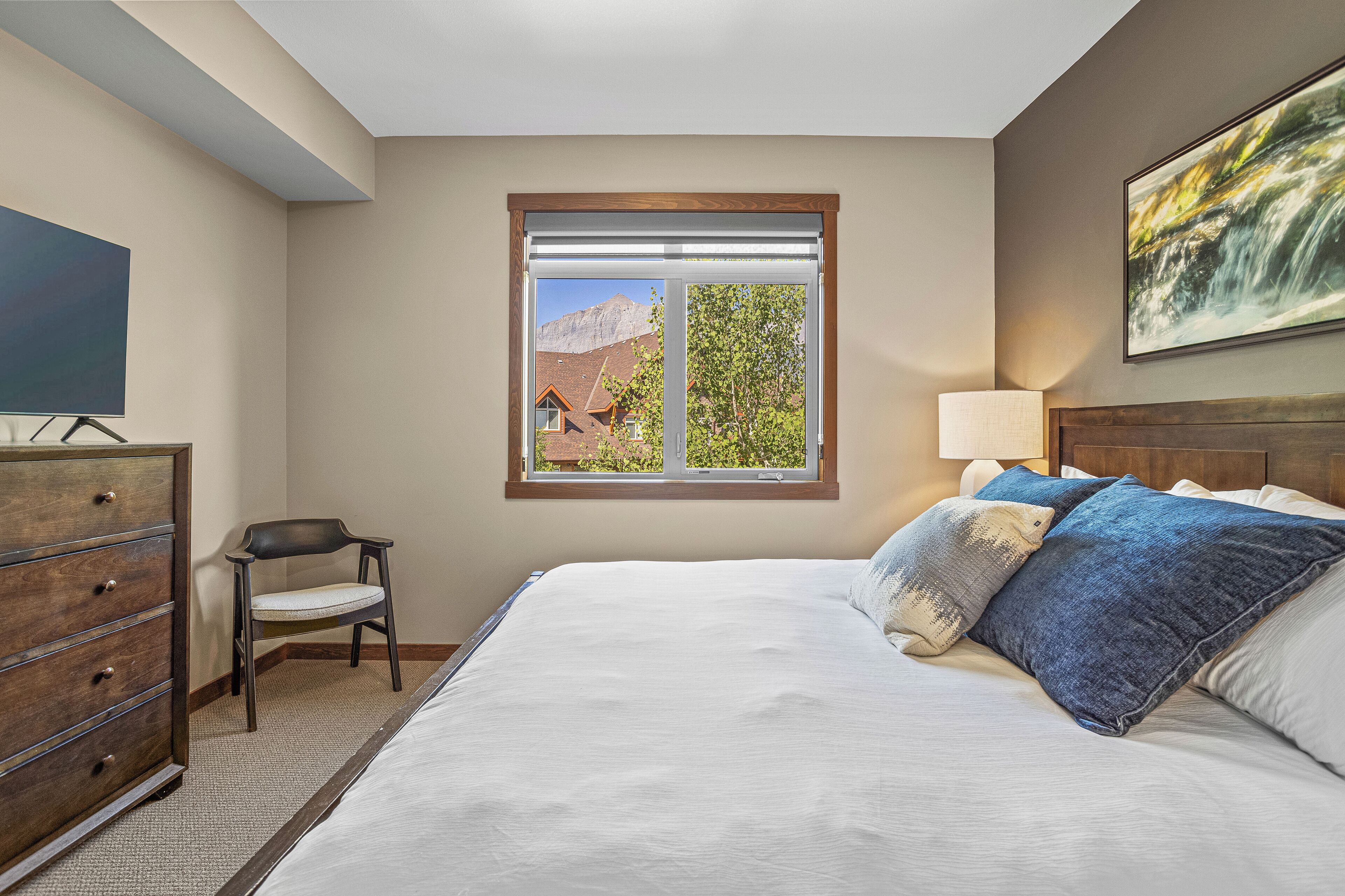 Une chambre avec un grand lit, une commode, une chaise, une fenêtre donnant sur la montagne et un tableau accroché au mur.