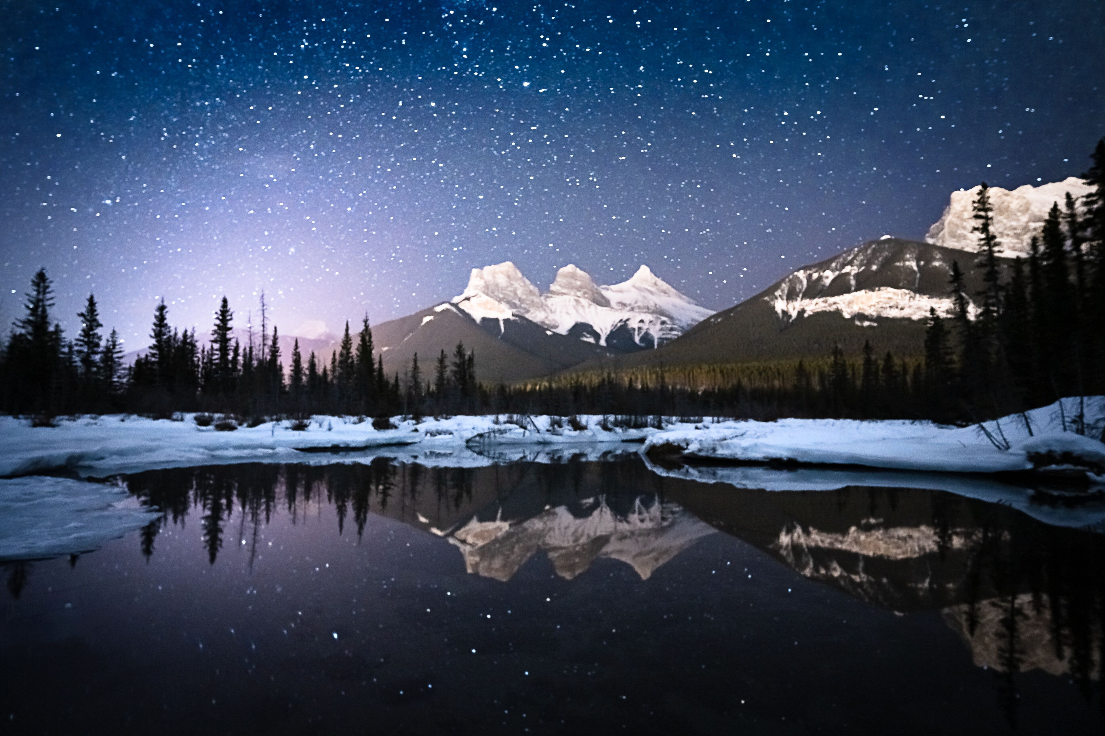 Céu estrelado sobre uma cordilheira refletida num lago calmo, rodeado por árvores.
