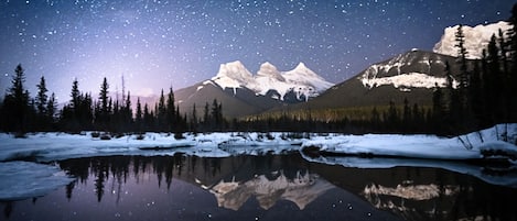 Céu estrelado sobre uma cordilheira refletida num lago calmo, rodeado por árvores.