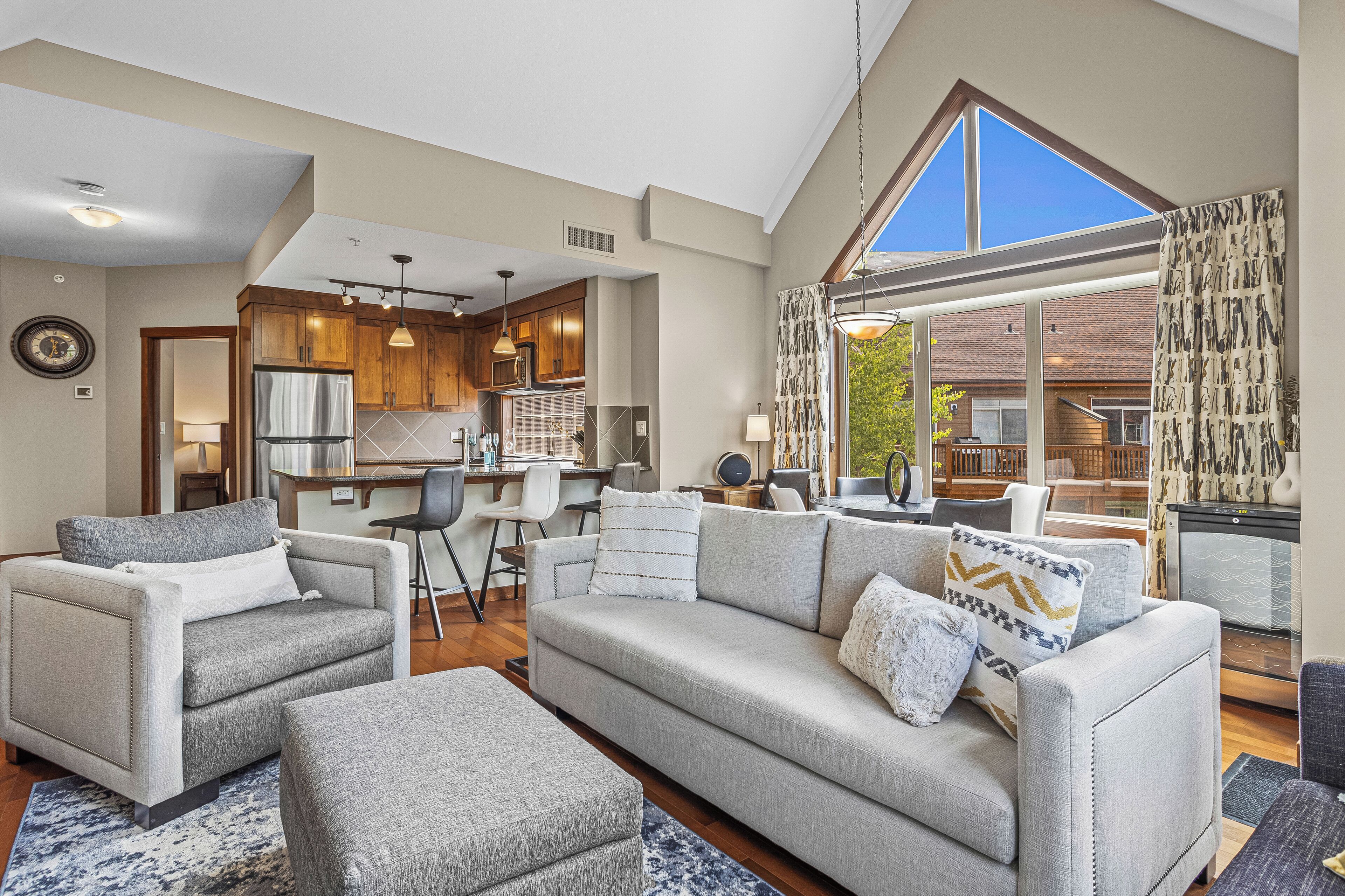 A spacious living area with a grey sofa set, a dining table, and a kitchen in the background.