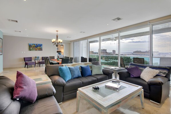 A spacious living room with a large window, a sofa set, a coffee table, and a dining area.