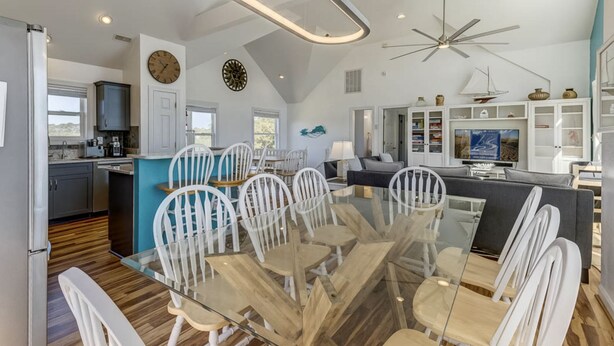 A spacious kitchen with modern appliances, a dining area with a glass table and white chairs, and a living area with a large TV and decorative items. - A Beach Vacation destination. EV friendly. (Corolla)