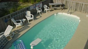 A rectangular swimming pool with clear water, surrounded by a wooden deck. There are several white plastic chairs and a table on the deck. - A Beach Vacation destination. EV friendly. (Corolla)