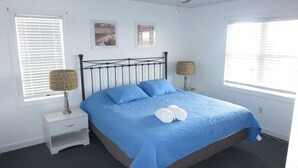 A neatly made bed with blue bedding and two pillows, a bedside table with a lamp, and a window with blinds. - A Beach Vacation destination. EV friendly. (Corolla)