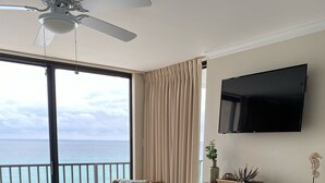 Une chambre avec un lit, une chaise en osier, une table avec des plantes, une télévision et un balcon donnant sur la mer.