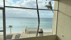 Une vue sur la plage depuis une chambre dotée d’une grande fenêtre, d’une clôture blanche et d’un palmier.