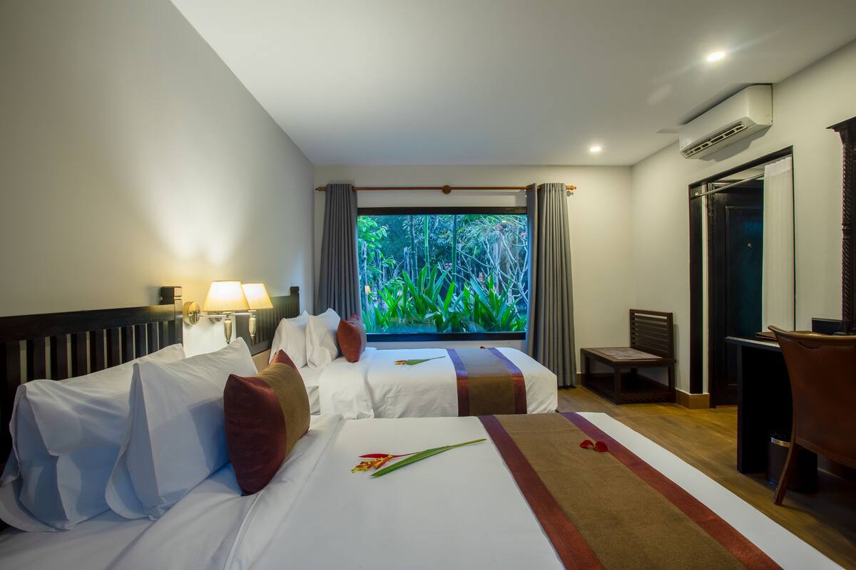 Une chambre d’hôtel avec deux lits, un bureau, une chaise et une vue sur la verdure par la fenêtre.