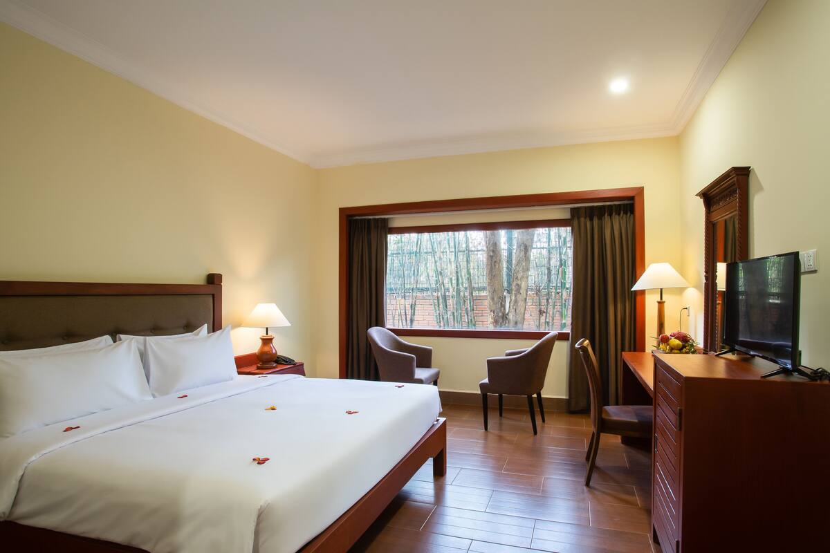 Une chambre d’hôtel avec un grand lit, deux fauteuils, un bureau avec une télévision et une fenêtre avec des rideaux.