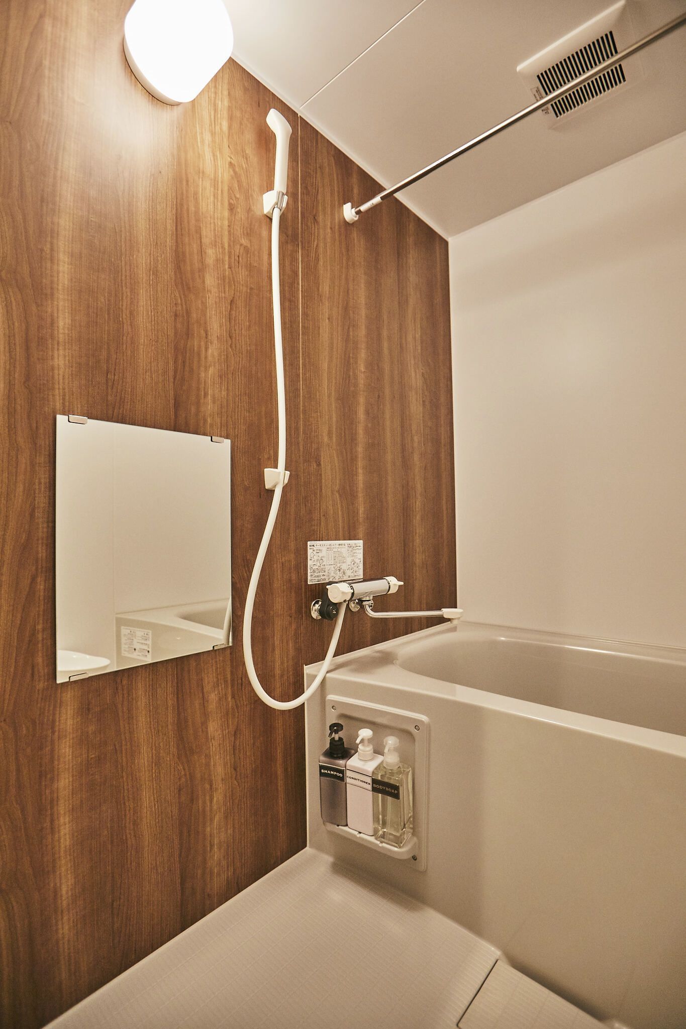 A modern bathroom with a wooden wall, a white bathtub, a handheld showerhead, a mirror, and a wall-mounted soap dispenser.