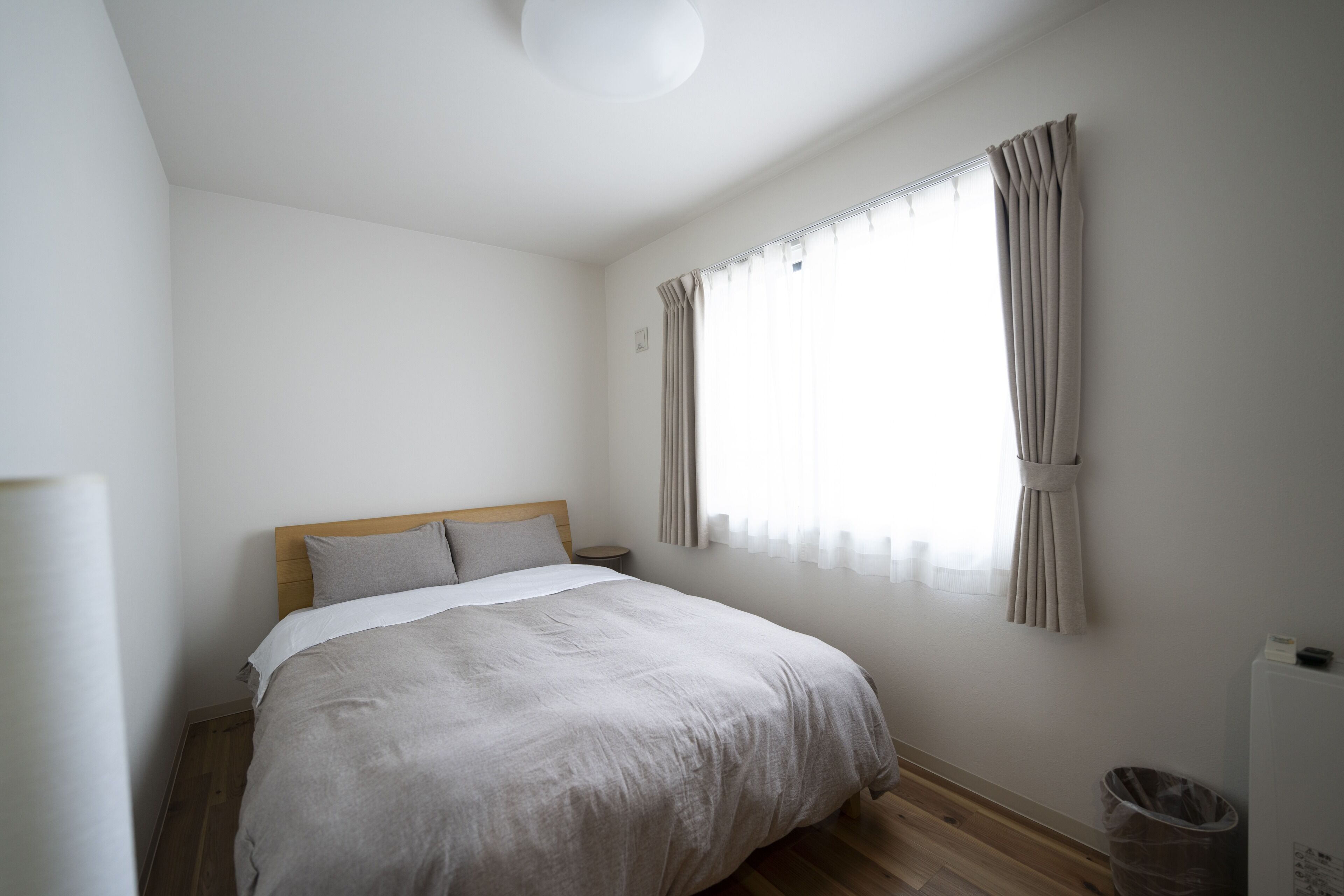 A bedroom with a bed, a window with curtains, a nightstand, and a trash bin.