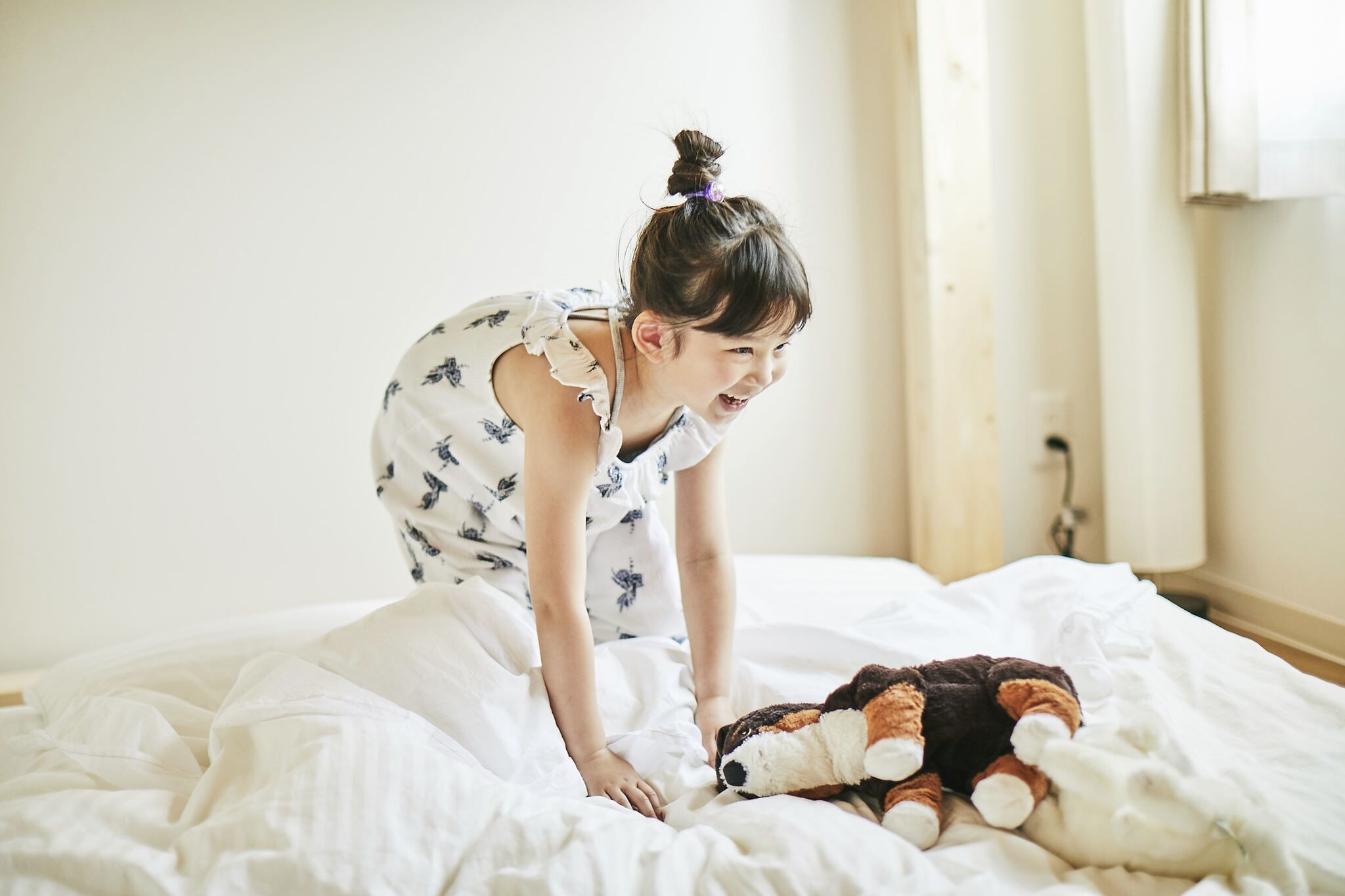 Un enfant joue avec un jouet en peluche sur un lit, dans une pièce aux murs blancs et dotée d’une fenêtre.