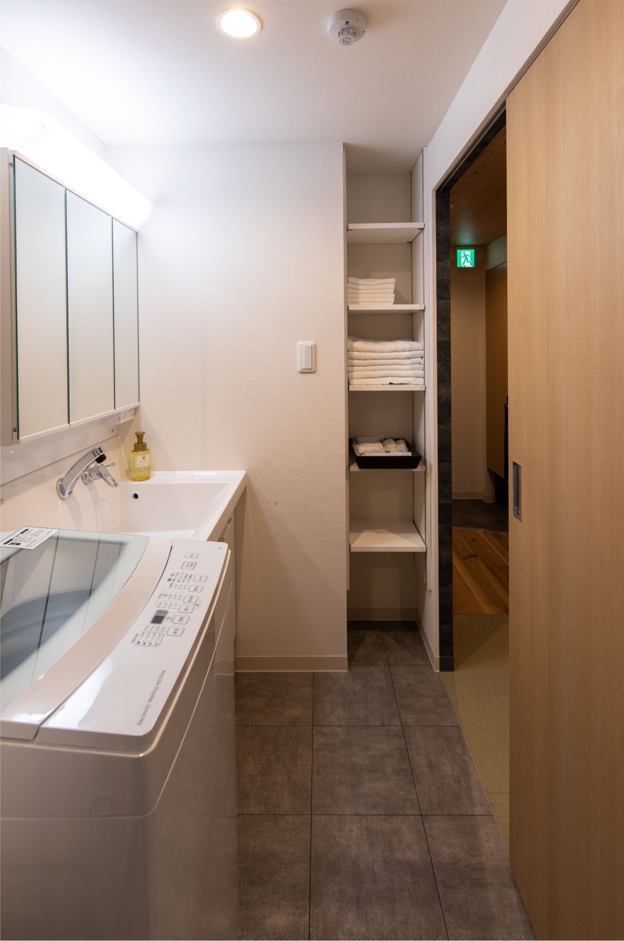 Un baño moderno con lavadora incorporada, un lavabo y una ducha. Una puerta de madera da a otra habitación.