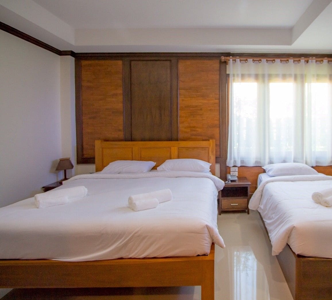 Camera d'albergo con due letti, testiere in legno, lenzuola bianche, un comodino con una lampada e una finestra con tende leggere.