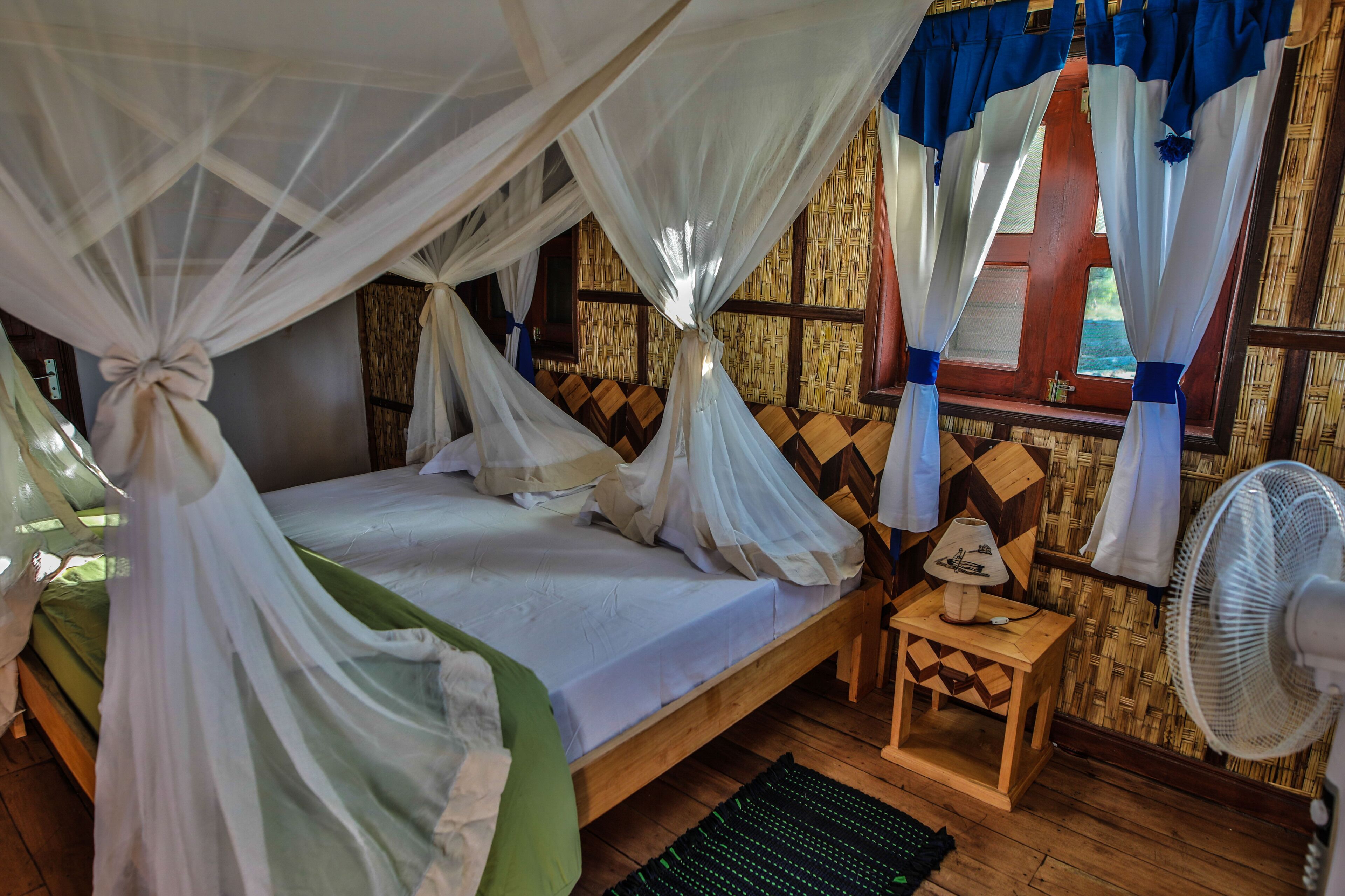Una habitación con una cama bajo un dosel, una pequeña mesa de madera con una lámpara, un ventilador y una ventana con cortinas.