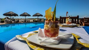 Mesa junto a la piscina con una bebida tropical y un plato, rodeada de sombrillas y un cielo azul despejado.