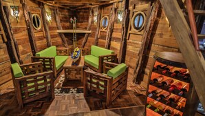 Interior de una cabaña de madera con una mesa de comedor de madera, cuatro sillas de madera con cojines verdes, una estantería para vinos con botellas y ventanas circulares.