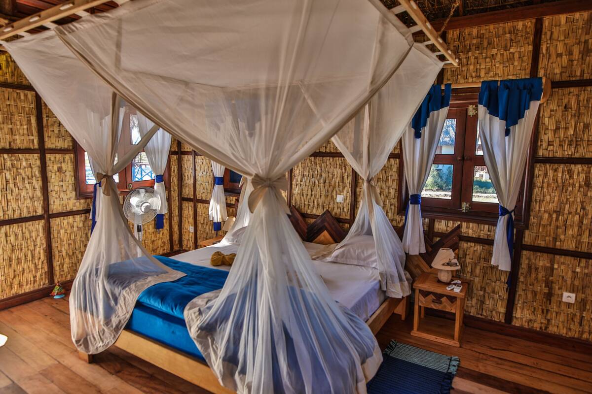 A room with a bed covered by a mosquito net, wooden walls, and a window with curtains.