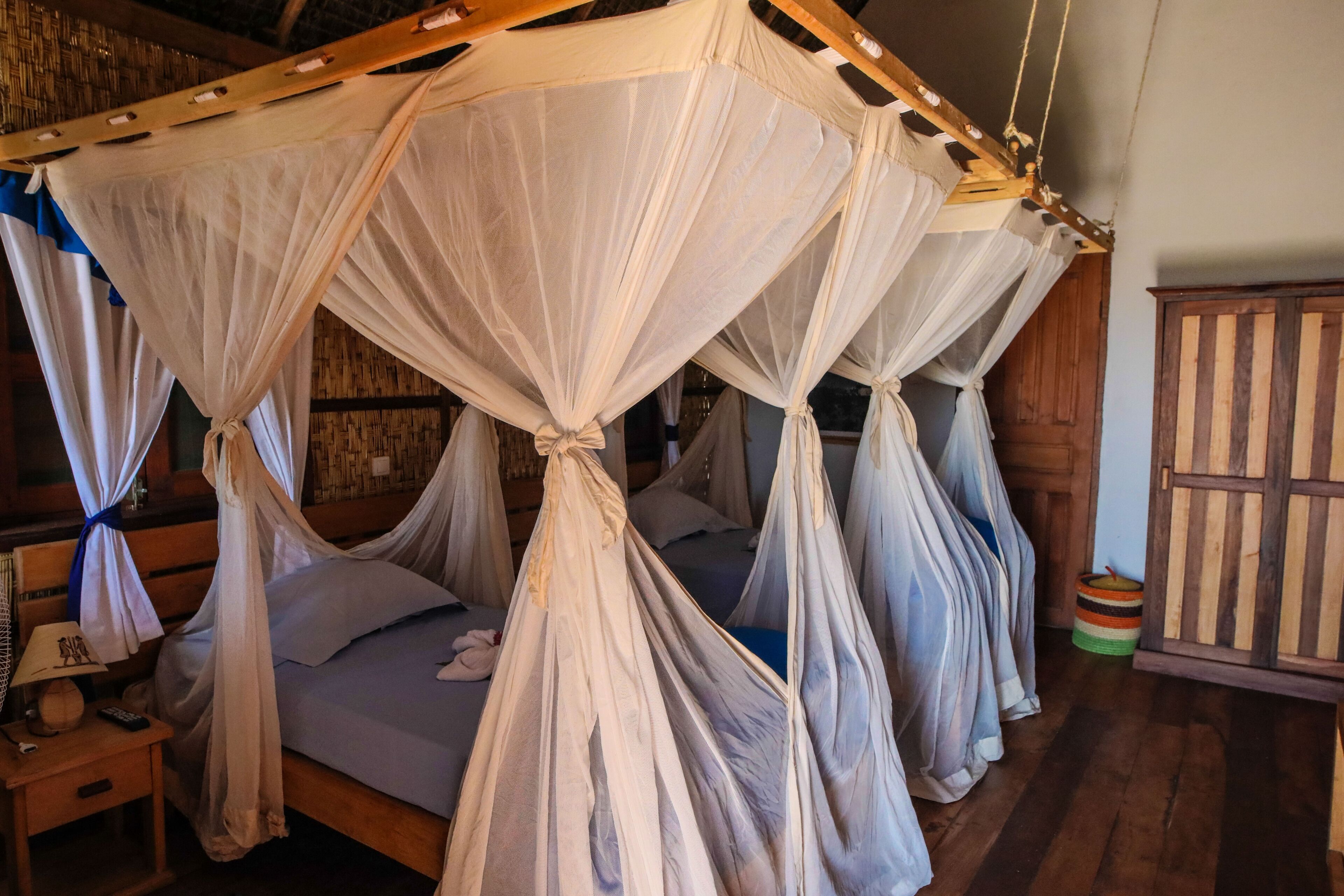 Une chambre avec quatre lits, chacun surmonté d’un dais. Il y a une tête de lit en bois sur le côté droit, une table de chevet avec une lampe et une petite table sur laquelle est posé un cadre photo.