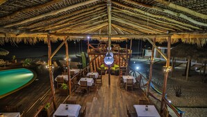 Un restaurante al aire libre con mesas y sillas de madera, techo de paja y vista a la playa.
