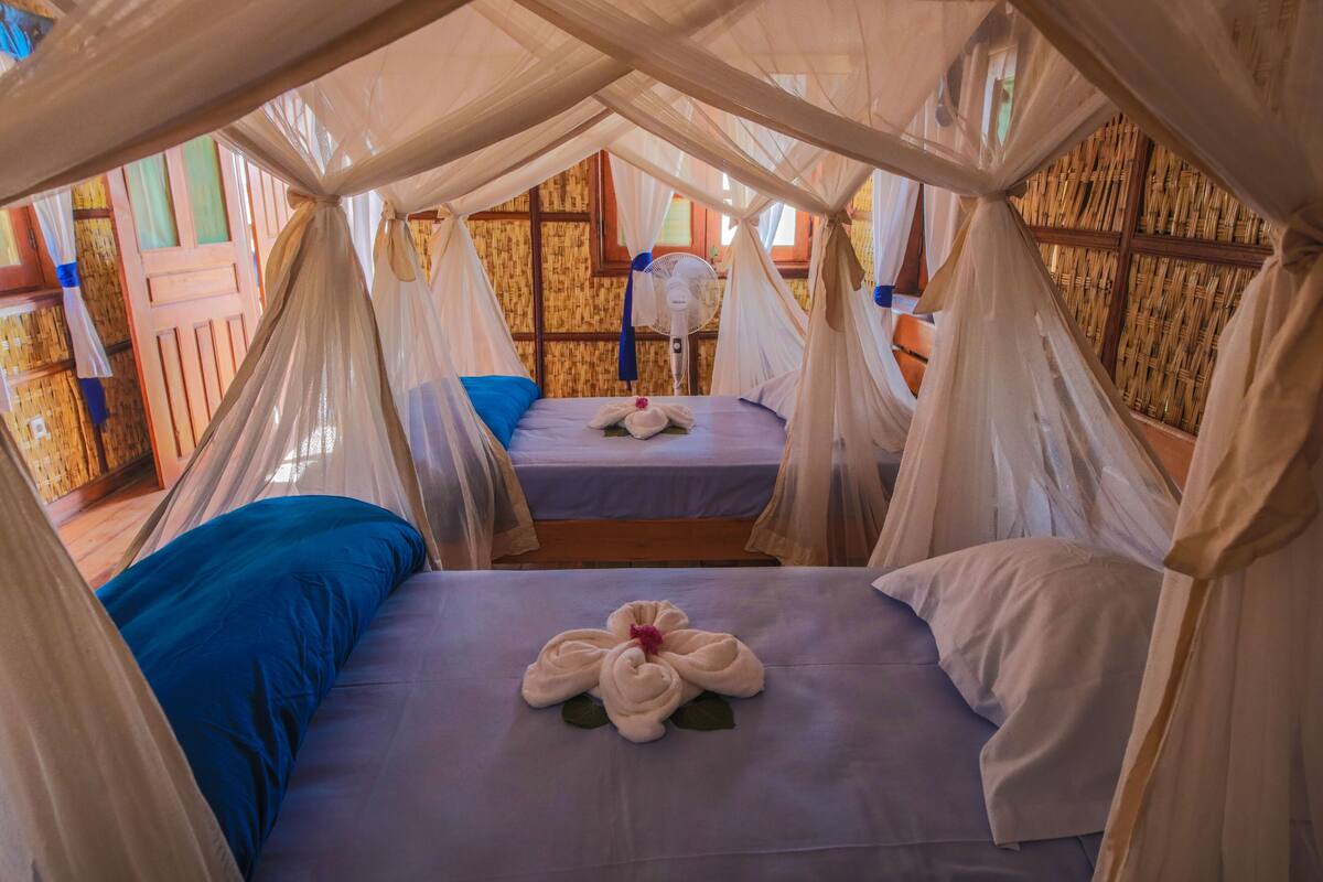 A room with two beds, each with a canopy. There is a fan on one bed and a flower arrangement on the other.