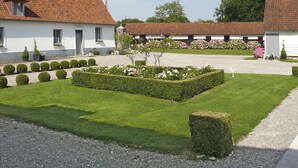 Une cour bien entretenue, avec des haies soigneusement taillées, un petit arbre et un chemin de gravier menant à un bâtiment blanc surmonté d’un toit en tuiles rouges.
