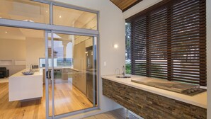 A modern kitchen with a glass door leading to a dining area, a built-in brick backsplash, and a window with horizontal blinds. - Juniper Metricup House - luxury rural retreat (Wilyabrup)