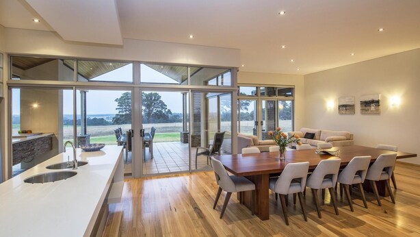 A modern kitchen with a large island, a dining area with a wooden table and chairs, and large sliding glass doors leading to an outdoor patio. - Juniper Metricup House - luxury rural retreat (Wilyabrup)
