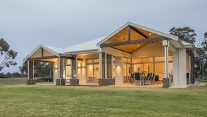 A modern house with a large patio, a dining area, and a kitchen visible through large windows. - Juniper Metricup House - luxury rural retreat (Wilyabrup)