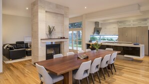 A modern kitchen with a central island, a dining area with a wooden table and chairs, and a living area with a sofa and fireplace. - Juniper Metricup House - luxury rural retreat (Wilyabrup)