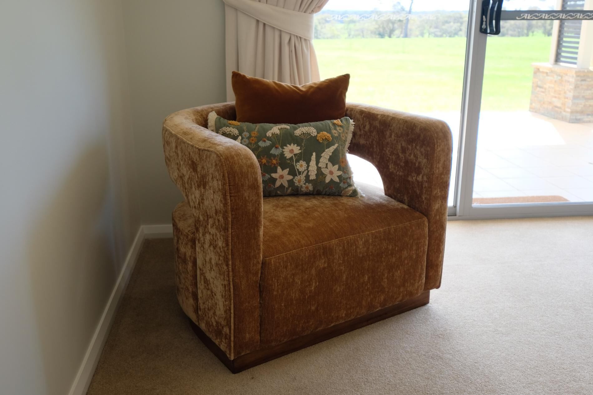 A brown armchair with a floral pillow in a room with a view of a grassy field through a glass door.