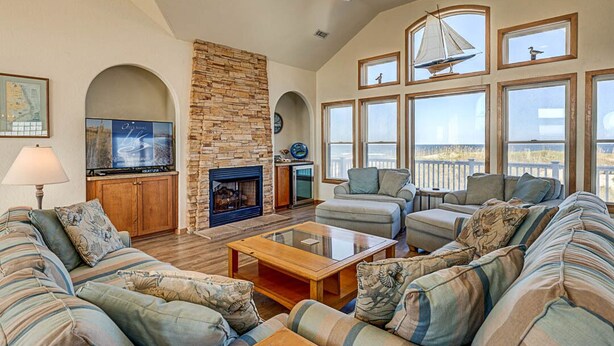 A spacious living room with a stone fireplace, comfortable seating, large windows, and a ceiling fan. - The Lodge | Oceanfront | Private Pool, Hot Tub | Nags Head (Nags Head)