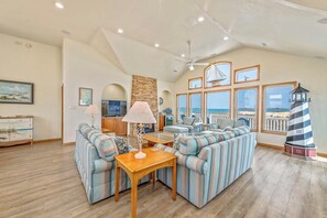 A spacious living room with a large sectional sofa, a wooden coffee table, and a lamp. There are multiple windows offering a view of the beach. - The Lodge | Oceanfront | Private Pool, Hot Tub | Nags Head (Nags Head)