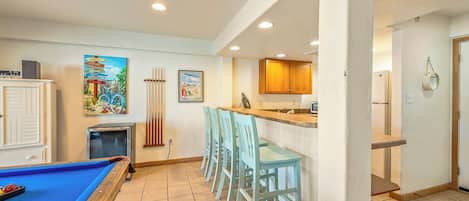 A pool table with a blue felt surface, a bar area with a counter and stools, and a kitchen with wooden cabinets.